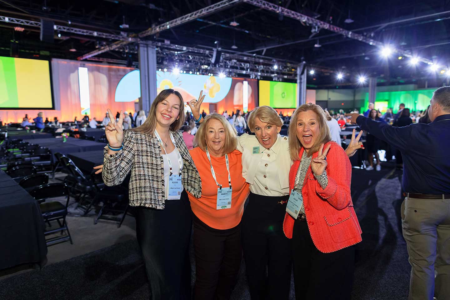 Four women including Cathy burns smiling and posing with peace signs at an indoor event with bright lighting and a large screen in the background.