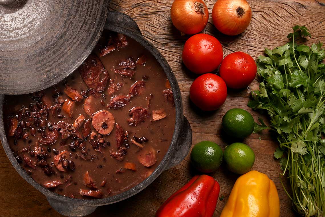 A clay pot with a stew of black beans and sausage, surrounded by fresh vegetables on a wooden surface.