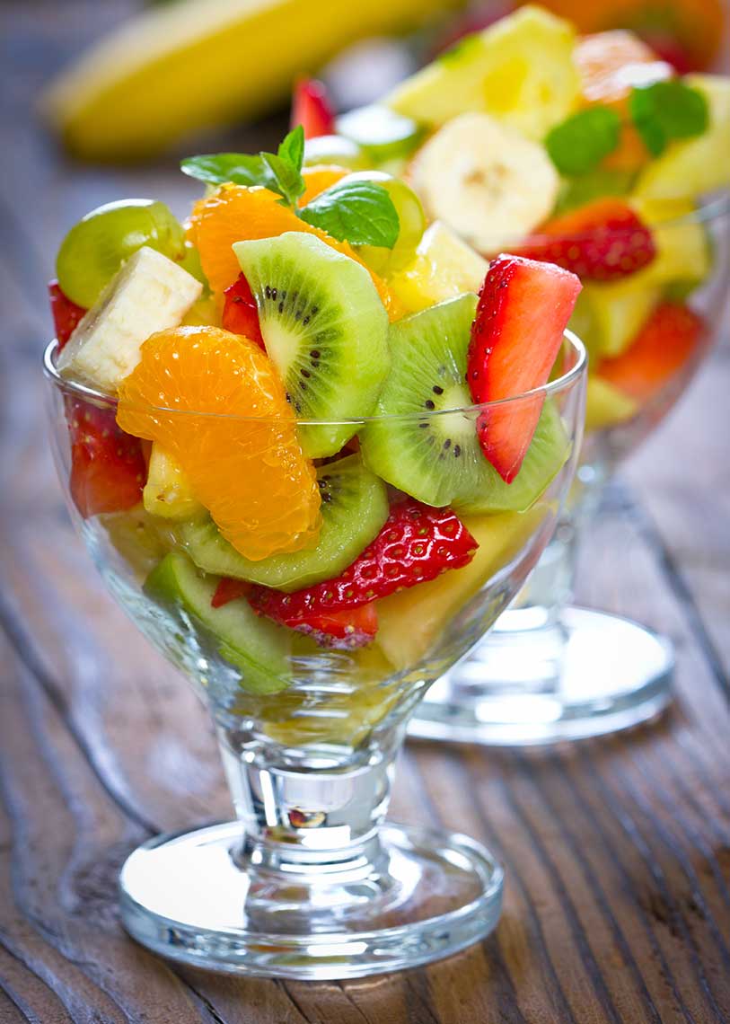 Fruit salad in a glass cup with kiwi, mandarin, strawberries, bananas, and grapes, garnished with mint leaves.