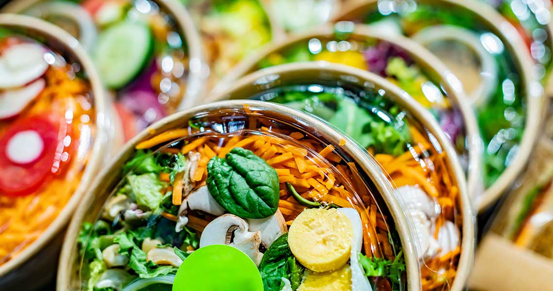Close-up of several salad bowls covered with clear lids, showcasing spinach, carrots, mushrooms, and sliced egg.