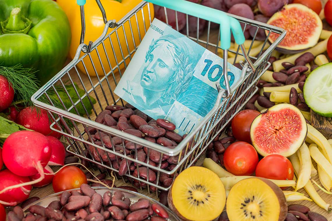 A shopping basket with red kidney beans and a 100 Brazilian real banknote, surrounded by fresh fruits and vegetables.