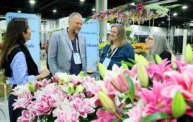 Four people talking at a floral exhibition with pink and white lilies in the foreground.