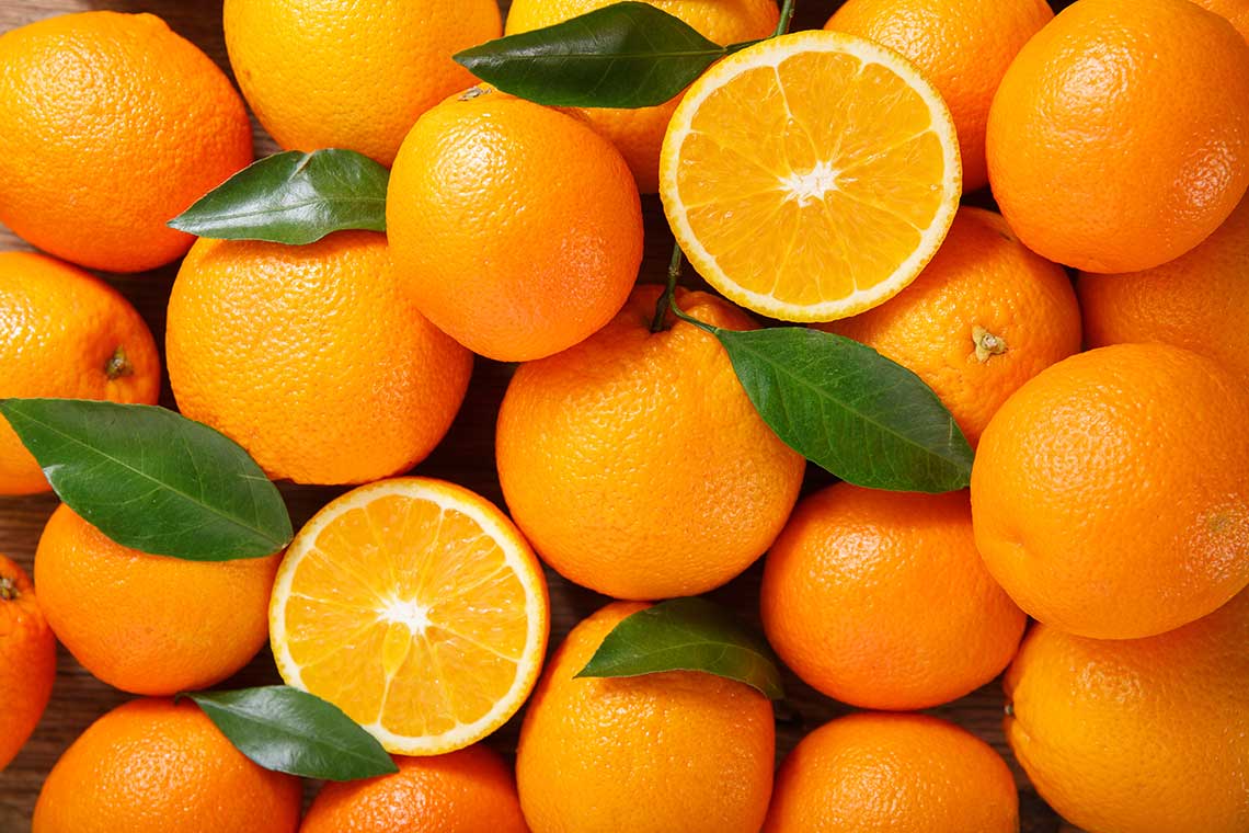 A pile of fresh oranges with two cut in half, showing the interior, and some green leaves scattered among them.